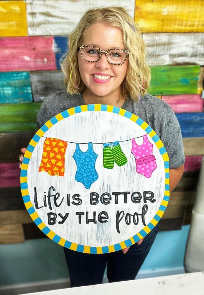 Tamara holding a Door Hanger that reads "Life is Better by the Pool" with swimsuits on a clothesline