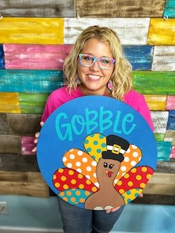 Tamara holding the Gobble Turkey door hanger in front of her pallet wall