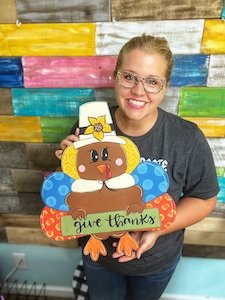 Tamara Bennett holding a painted turkey door hanger that reads "give thanks"
