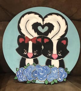 two skunks on a round door hanger with flowers tails together to form a heart
