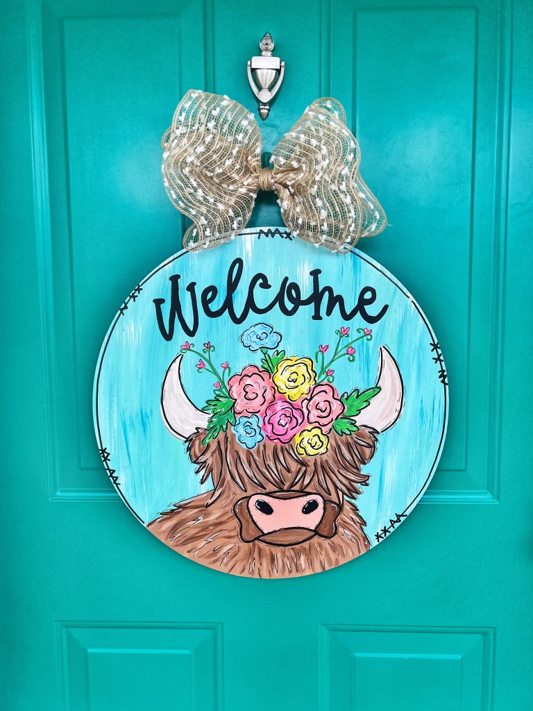 Door hanger with highland cow with flowers on head that says "welcome" across the top. burlap bow on top of door hanger that is hung on teal door