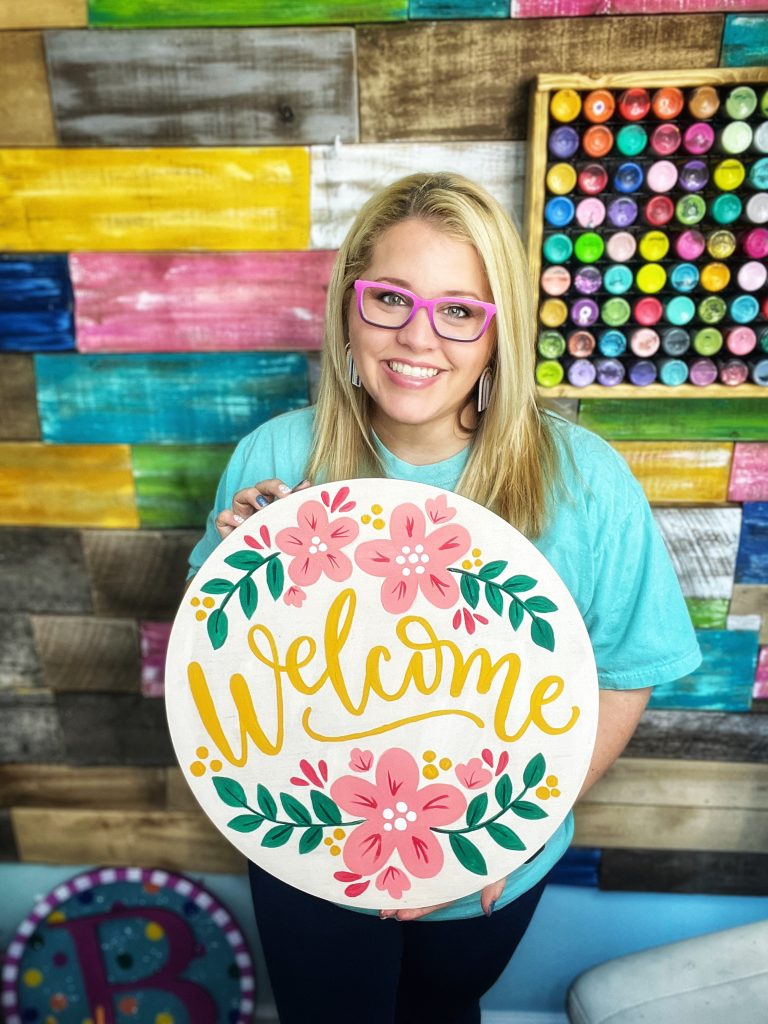 Tamara holding a round door hanger that says "welcome" with pink flowers and greenery