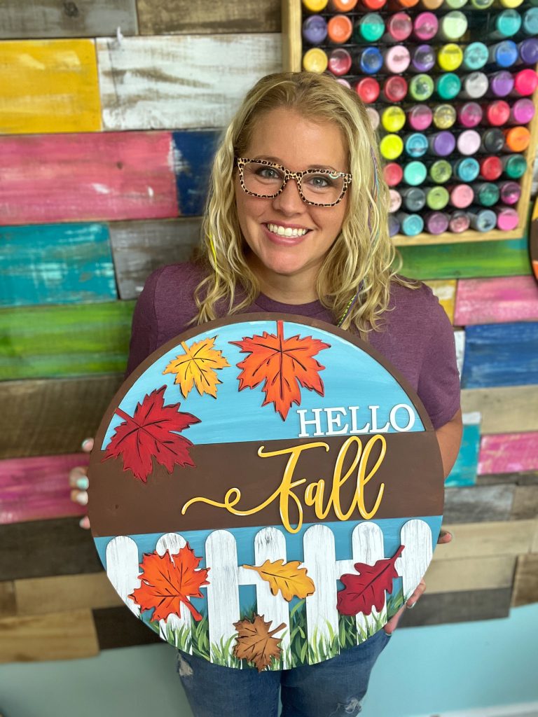 woman holding a door hanger that says "hello fall" with white picket fence and leaves