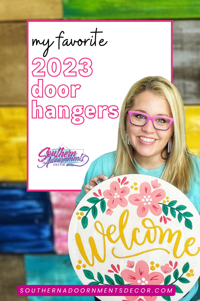 Tamara holding a door hanger that says "welcome" and has flowers in the background on a painted wood pallet background. Picture reads "my favorite 2023 door hangers"