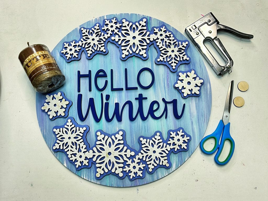 door hanger that says "hello winter" with snowflakes on it, laying on a surface with jute chord, stapler, scissors, and small wooden discs